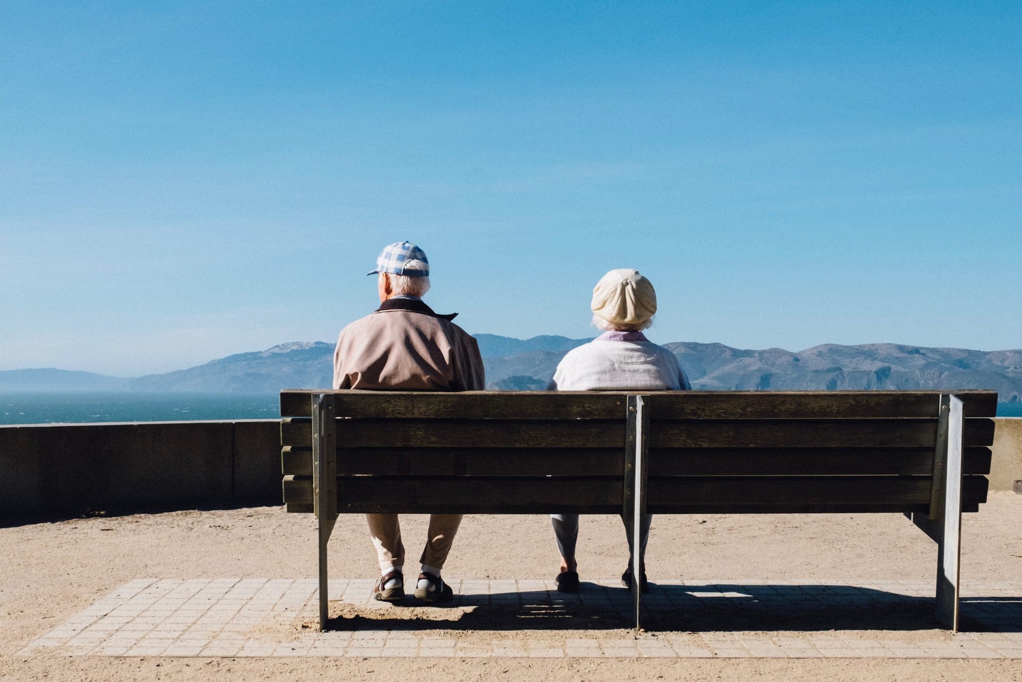 Two people sitting on a bench looking at the view of the sea and hills. Aster Care offers live-in care services for those with long-term conditions. Click to find out how we provide care today.