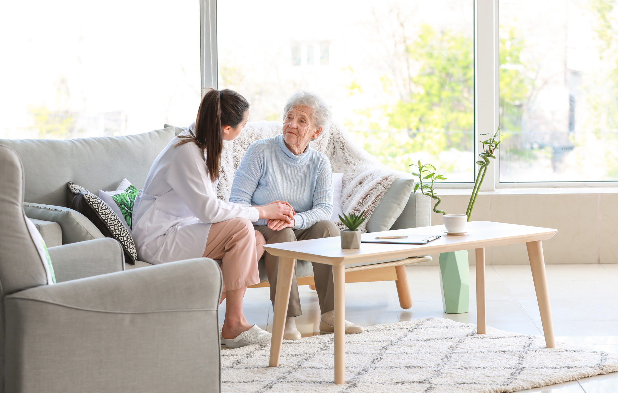 A live-in carer, in a bright, airy room, sat on a grey sofa with her client, comforting her. Live-in Care with Aster Care is an affordable alternative to nursing home care.